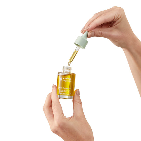 A hand holding a 30ml glass dropper bottle of endota Phyto Renewal Oil in one hand, while the other hand is holding the dropper cap. The bottle is prominently displayed with its label facing forward. The background is white.