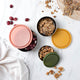 3 round stainless steel containers with earthy coloured lids styled on a marble counter with snacks.