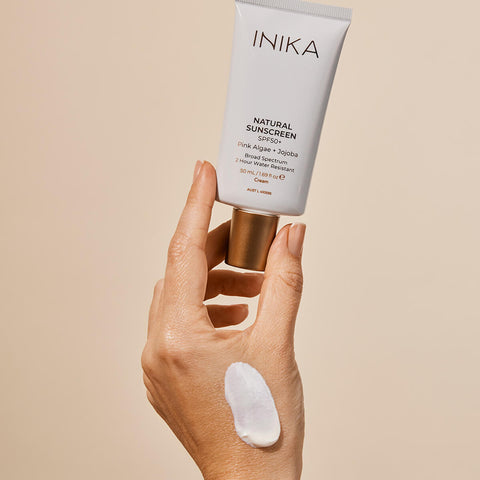 Hand holding tube of certified organic sunscreen with SPF50+, pink algae and jojoba, with a swatch of the product on the hand.