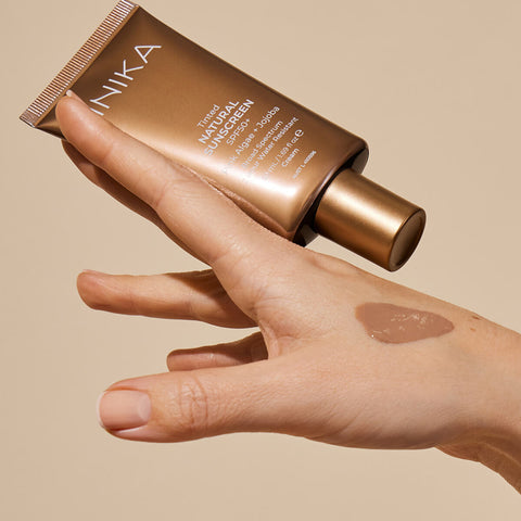 Hand holding tube of certified organic tinted sunscreen with SPF50+, pink algae and jojoba, with a swatch of the product on the hand.