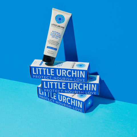 100ml bottle of Little Urchin Natural Clear Zinc Sunscreen SPF 50+ balanced on a stack of its boxes on a blue surface. The sunscreen is a hydrating, vegan formula that provides UVA and UVN broad spectrum protection from the sun while also being 4 hours water-resistant. Packaged in a cream tube with minimal blue and black text on the label.