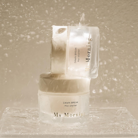 45g jar of Ms Morning Dawn Break Moi-Starter, a 3-in-1 product that serves as an anti-aging serum, moisturiser, and primer, combining the powerful hydrating and anti-aging benefits of hyaluronic acid and hydrolysed collagen to deliver exceptional skincare results. There's 2 jars balanced on top of each other in a rainy scene. Packaged in a frosted glass jar with gold text.