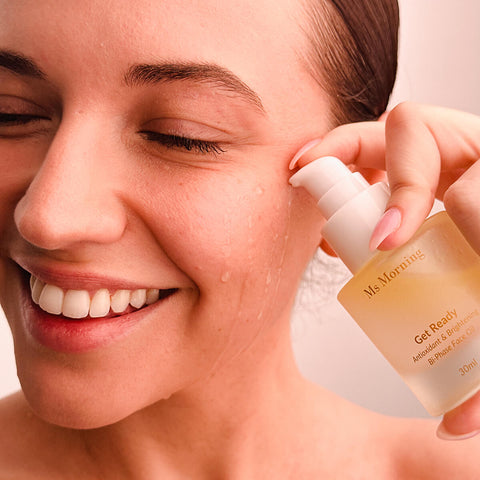 A woman applying Ms Morning Get Ready Antioxidant & Brightening Bi-Phase Face Oil to her face. The oil is infused with organic antioxidants and brightening ingredients, designed to protect and prep the skin. The product is packaged in a frosted glass bottle with a gold cap.