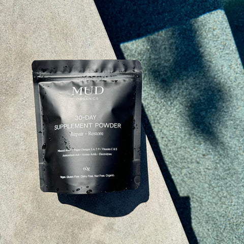 Mud Organics 30-Day Supplement Powder placed next to a pool. The powder features 70+ trace minerals, zinc, magnesium, vitamin C and E, antioxidants, and omegas 3, 6, 7, and 9 that supports energy, immunity, and improved cognitive function. Packaged in a resealable black plastic bag.