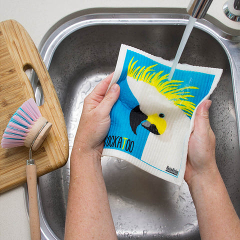 Hands holding a compostable sponge cloth with a cockatoo design in a sink.