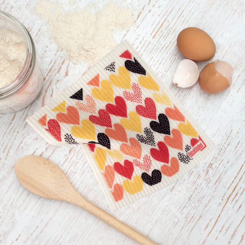 RetroKitchen Compostable Sponge Cloth, a super absorbent, machine washable and compostable dish cloth with a heart design on a bench with a wooden spoon, flour, and eggs.