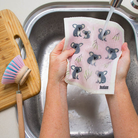 Hands holding a compostable sponge cloth with a koala design in a sink.