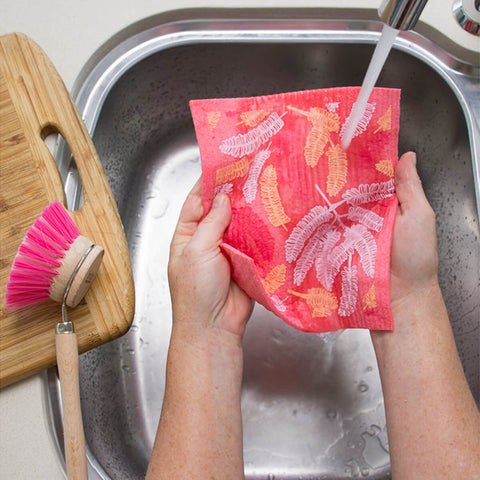 Hands holding a compostable sponge cloth with a poinciana design in a sink.