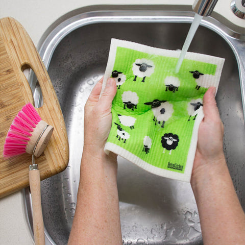 Hands holding a compostable sponge cloth with a sheep design in a sink.