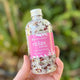 Hand holding a 300g glass bottle of The Physic Garden Bath Soak in Relax against a green background. Packaged with a pink label.