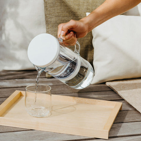 Hand pouring water into a glass on a wooden tray using a 2L waterdrop Lucy Filter Carafe water filtering jug made of robust, odourless and tasteless borosilicate glass, and features a minimal and sleek design.