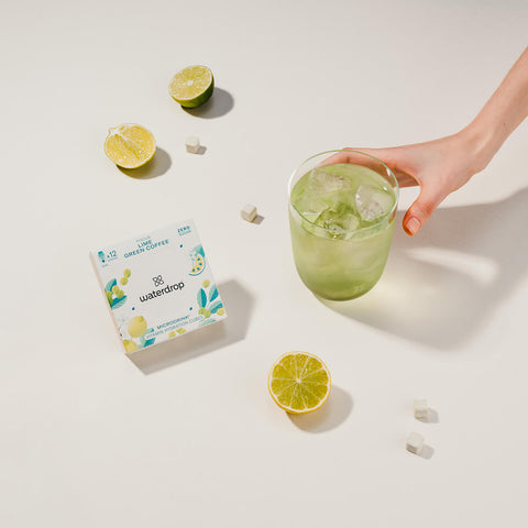 A hand holding a glass of lime and green coffee flavoured water with ice, next to a white box of 12 waterdrop Microdrink cubes in the Focus flavour. The box displays a lime and green coffee flavour, with blue, green and yellow lime and coffee bean illustrations on the front. Scattered around the scene are small hydration cubes and limes on a light cream surface.