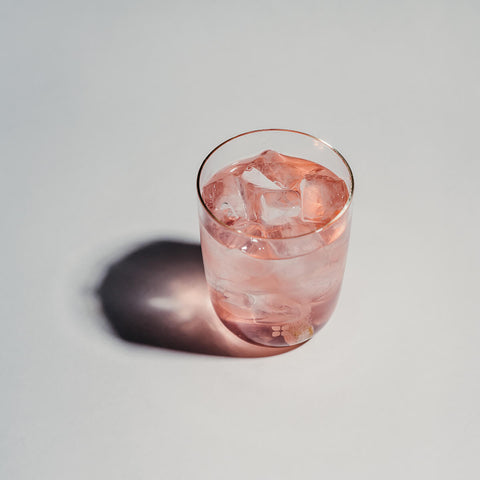 A glass of golden berry, peppermint and rosemary flavoured water with ice, and a waterdrop Microdrink cube in the Vibe flavour dissolving at the bottom. The water has a light pink colour.