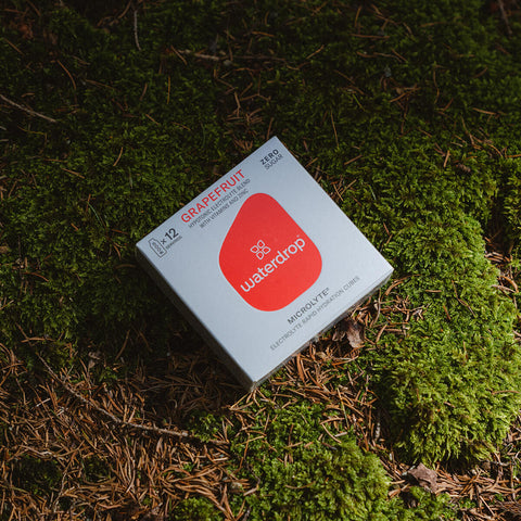 A box of 12 waterdrop Microlyte cubes in grapefruit flavour on a mossy surface. These sugar-free electrolyte cubes are made with natural fruit and plant extracts, providing a vitamin and zinc-enriched, flavoured drink. Each cube dissolves to create 400-600ml of a refreshing beverage designed to support you both during and post-exercise. The product comes in a silver box with a bright red logo in the middle.